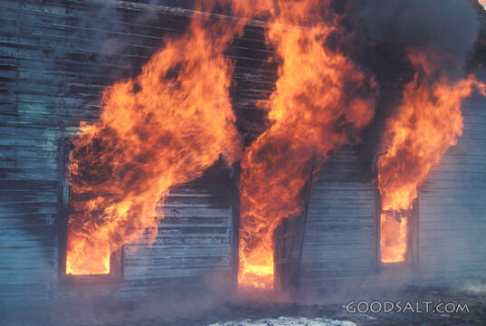 Flames bursting through the door and windows of an old building.