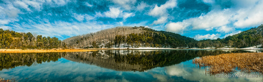 Snow and trees on the other side of a lake are reflected in its waters.