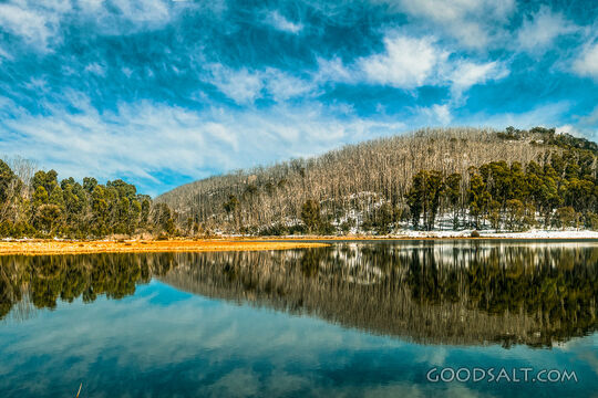Snow and trees on the other side of a lake are reflected in its waters.