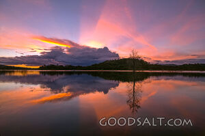 Dramatic sunset and storm cloud over still lake waters.