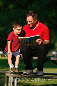 Man reading Bible to little boy in park in summer.