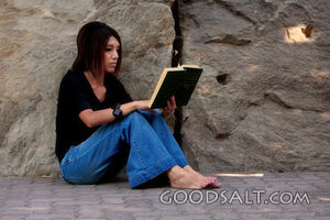 Woman reading, leaning against stone wall.